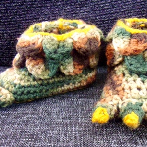 May include: A pair of crocheted slippers with a camouflage pattern and yellow toes. The slippers are made with brown, green, and beige yarn.