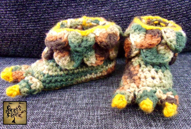 May include: A pair of crocheted slippers with a camouflage pattern and yellow toes. The slippers are made with brown, green, and beige yarn.