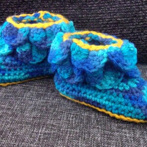May include: Blue and yellow crocheted slippers shaped like dragon claws. The slippers have a scalloped edge and are made with a textured yarn.