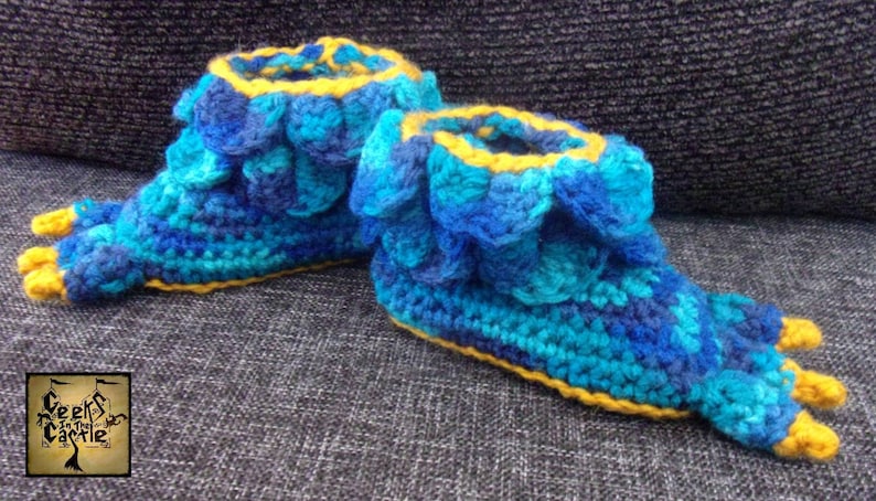 May include: Blue and yellow crocheted slippers shaped like dragon claws. The slippers have a scalloped edge and are made with a textured yarn.