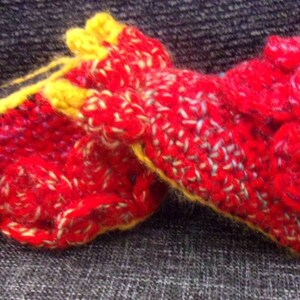 May include: A pair of red and yellow crocheted slippers with a textured design. The slippers are on a gray fabric surface.