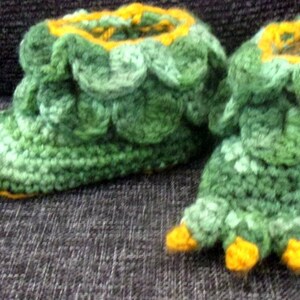 May include: A pair of green and yellow crocheted slippers shaped like a dinosaur foot. The slippers have a yellow rim and toes.
