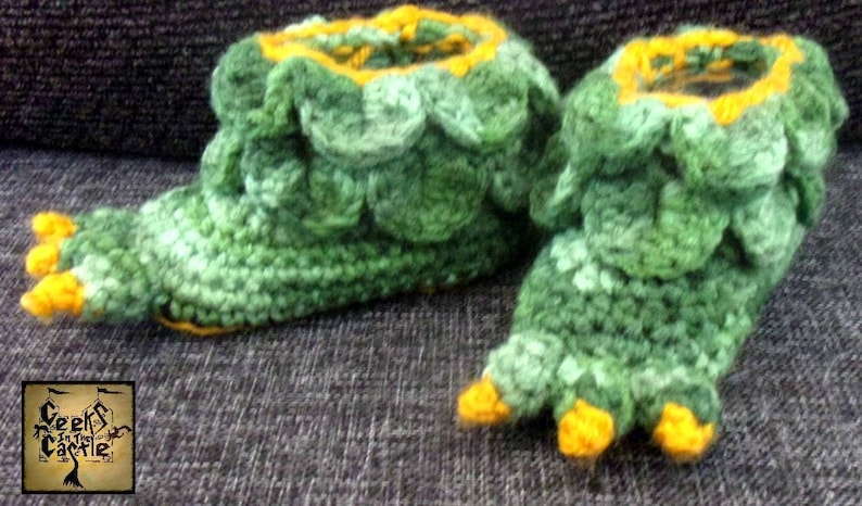 May include: A pair of green and yellow crocheted slippers shaped like a dinosaur foot. The slippers have a yellow rim and toes.