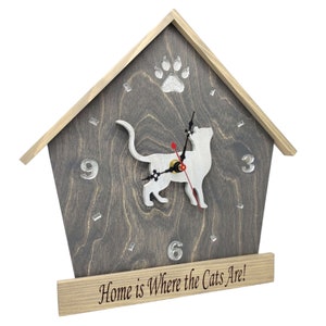 May include: A wooden house-shaped clock with a white cat silhouette and a paw print design. The clock face has Roman numerals and the text "Home is Where the Cats Are!" is printed on the base.