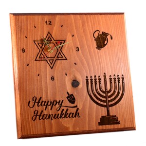 May include: Square wooden clock with a light brown finish. The clock face features a Star of David, a menorah, and the words "Happy Hanukkah." The clock hands are gold, and the numbers are etched into the wood.