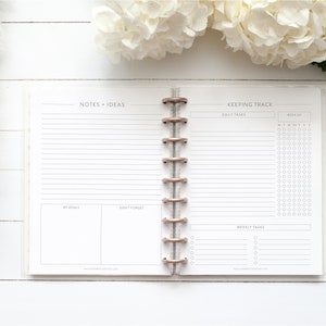 May include: A white planner with a clear plastic cover. The planner is open to a page with the headings "Notes + Ideas" and "Keeping Track". The left page has lined space for notes and a section for "My Goals" and "Don't Forget". The right page has lined space for "Daily Tasks" and a grid for tracking tasks by day of the week. There is also a section for "Weekly Tasks".