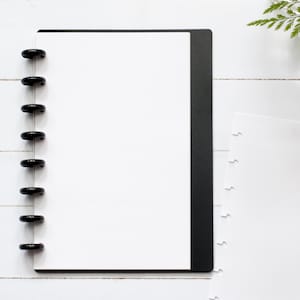 May include: A black and white notebook with a black cover and a black binding. The notebook has a white interior with a dotted pattern. The notebook is lying flat on a white surface.