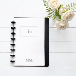 May include: A white to-do list notebook with a black spiral binding. The notebook has the text "TO DO LIST" printed on the front cover. The notebook is on a white wooden surface with a vase of white roses.
