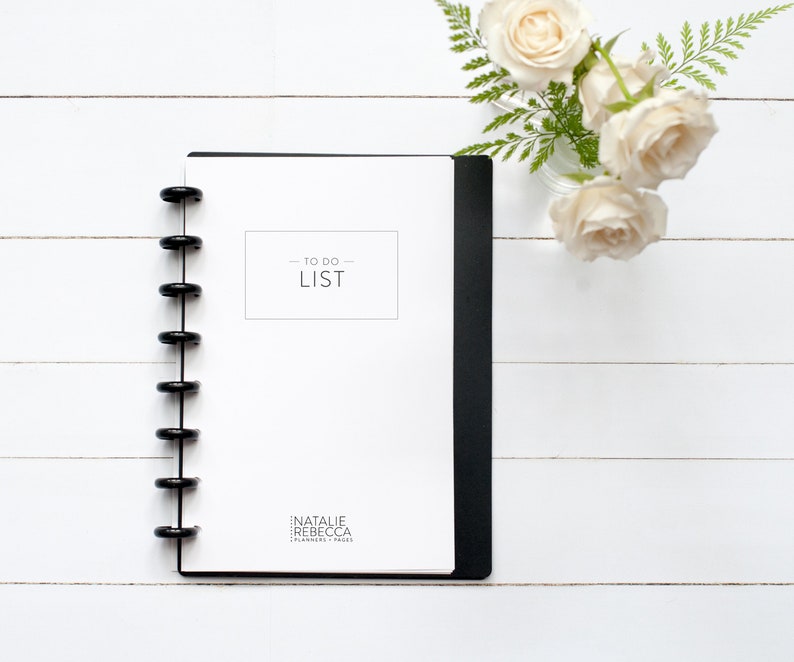 May include: A white to-do list notebook with a black spiral binding. The notebook has the text "TO DO LIST" printed on the front cover. The notebook is on a white wooden surface with a vase of white roses.