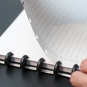 May include: A white notebook with black rings and lined pages. The top page is being turned over to reveal the next page.