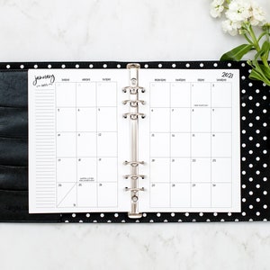 May include: A black and white polka dot planner with a silver ring binder. The planner is open to January 2021 and has a white background with black text. The planner is on a white marble surface.