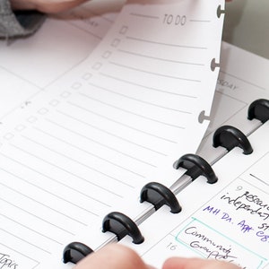 May include: A close-up of a black and silver planner with a white page turned to the "TO DO" section. The page has a checklist with several empty boxes. The planner has a black ring binding system.