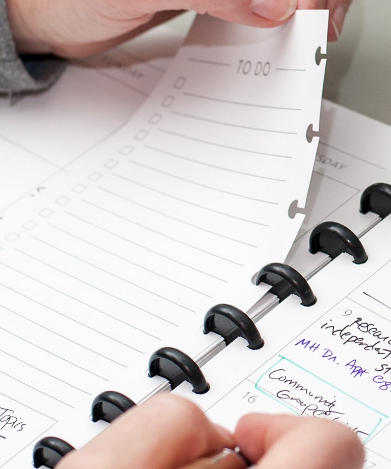 May include: A close-up of a black and silver planner with a white page turned to the "TO DO" section. The page has a checklist with several empty boxes. The planner has a black ring binding system.