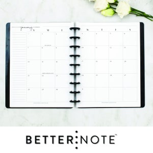 May include: A black and white monthly calendar planner with the month of January displayed. The planner has a black cover and a silver coil binding. The planner is open to the month of January and the days of the week are labeled at the top of the page. The planner is on a white surface.