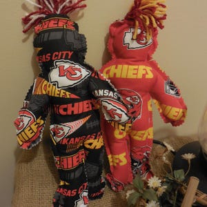 May include: Two handmade dolls, one black and one red, feature the Kansas City Chiefs logo and colors. Each doll has yarn hair in team colors. The dolls are standing on a burlap surface.