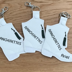 May include: Three white boot-shaped keychains with black embroidery. The keychains are decorated with a green tassel and the text "Rangerettes". One keychain has the name "Ariel" embroidered on it.