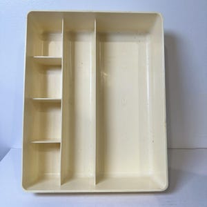 May include: A rectangular, off-white plastic utensil tray with multiple compartments. The tray has a large open area and several smaller sections for organizing silverware and kitchen tools. The tray is designed to fit inside a drawer.