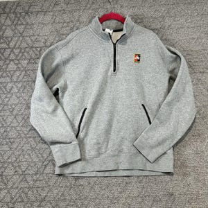 Peut inclure: Un sweat-shirt gris avec un col montant zippé et un petit logo sur la poitrine. Le sweat-shirt a des accents noirs sur les poches et la fermeture éclair. Le vêtement est suspendu à un cintre rose.