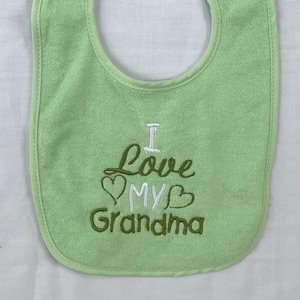 Puede incluir: Un babero de bebé de color verde claro con bordado blanco y verde que dice "Te quiero abuela".