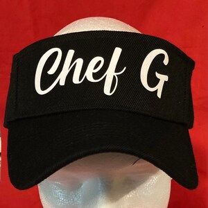 May include: Black chef hat with a white embroidered inscription that reads "Chef G".