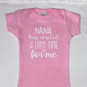 May include: Pink baby onesie with white text that reads "NANA has waited a long time for me" with a small heart symbol.