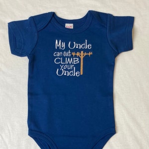 Puede incluir: Un body de bebé azul marino con bordado blanco que dice "Mi tío puede escalar más alto que tu tío" con una imagen bordada marrón de un poste de utilidad.