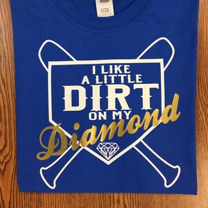 May include: Royal blue t-shirt with a white and gold graphic. The graphic features the text "I LIKE A LITTLE DIRT ON MY Diamond" with a baseball diamond and crossed bats design. The shirt is folded on a wooden surface.