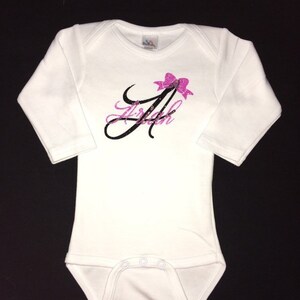 May include: White long-sleeve baby bodysuit with a black and pink glitter design of the letter "A" with a pink bow on top. The design says "Ariah" underneath the letter.