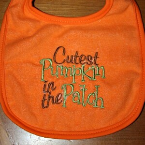 May include: Orange baby bib with the embroidered text "Cutest Pumpkin in the Patch" in brown, green, and white thread.