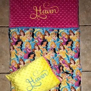 May include: A pink and yellow personalized nap mat with a princess pattern. The mat has a pillow with the name "Haven" embroidered on it. The mat is made of soft, comfortable fabric and is perfect for naps or playtime.