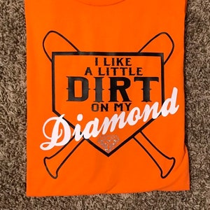 May include: Orange t-shirt with a baseball-themed graphic. The design includes a baseball diamond shape with crossed bats and the text "I LIKE A LITTLE DIRT ON MY DIAMOND" in black and white.