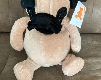 May include: A beige plush pug dog toy with black ears, nose, and eyes. The toy has a tag attached to its ear that says "26" and has a bear logo.