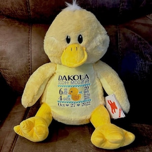 May include: A plush yellow duck stuffed animal with embroidered details. The duck has a white tuft of hair, black eyes, and a yellow beak and feet. The front of the duck has embroidered text and a graphic of a duckling.