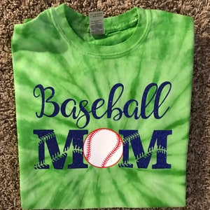 Puede incluir: Una camiseta verde teñida con la palabra "Baseball MOM" en letras azules brillantes. Un gráfico de béisbol está centrado en el diseño. La camiseta está doblada y parece ser de algodón.