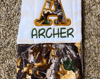Op de afbeelding: Een witte spuugdoek met een camouflagepatroon aan de onderkant. De bovenkant heeft een grote geborduurde letter A met een camouflagepatroon in de letter. De naam "ARCHER" is onder de letter geborduurd in groene draad.