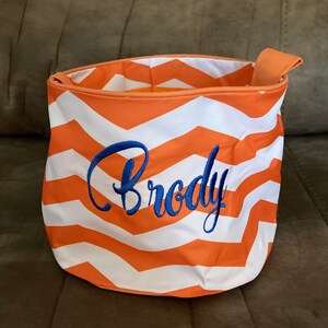 May include: Orange and white chevron patterned fabric storage bin with a blue embroidered name "Brody" on the front.