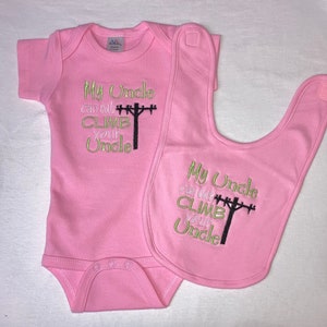 May include: Pink baby bodysuit and bib with embroidered text that reads "My Uncle can out climb your Uncle" with a black silhouette of a utility pole and power lines.