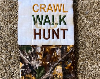 Op de afbeelding: Een witte baby spuugdoek met de woorden "Crawl Walk Hunt" geborduurd in bruin, groen en geel garen. De onderkant van de doek is gemaakt van camouflage stof.