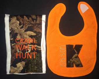 Op de afbeelding: Een witte slabbetje met een camouflagepatroon en de tekst "Crawl Walk Hunt" en een oranje slabbetje met een camouflageletter K en de tekst "Kason" in wit.