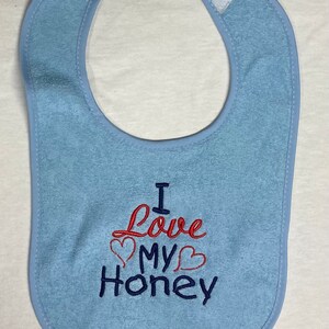 May include: A light blue baby bib with the embroidered text "I Love My Honey" in red and blue thread. The text includes two red hearts.