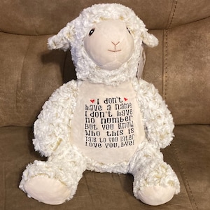 Op de afbeelding: Witte pluche lam knuffel met geborduurde tekst die luidt: "Ik heb geen naam Ik heb geen nummer maar je weet wie dit is Praat later met me Ik hou van je, doei!"