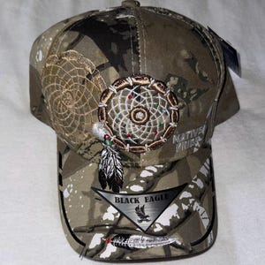 May include: Camouflage baseball cap with embroidered dreamcatcher and feather details. Features the text "Native Pride" and a "Black Eagle" emblem. The cap has a curved peak and a button on top.
