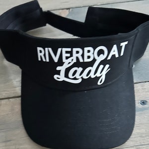 May include: Black visor with white text that reads "Riverboat Lady" in a cursive font.