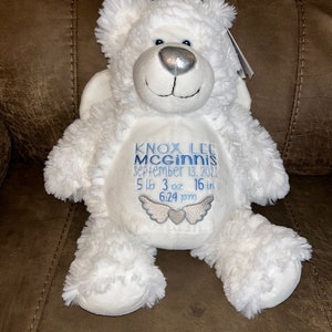 May include: A white plush teddy bear with embroidered details. The bear has a silver nose and blue eyes. The bear's belly features embroidered text in blue: "KNOX LEE MCGINNIS, September 13, 2022, 5 lb 3 oz 16 in, 6:24 pm" with angel wings.