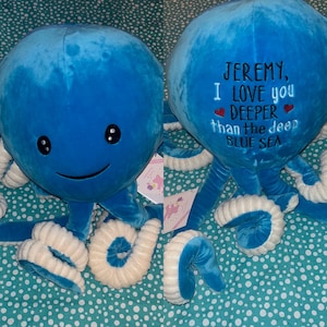 Op de afbeelding: Een blauwe pluche octopus met witte tentakels. De octopus heeft een glimlachend gezicht met twee zwarte ogen. De octopus is geborduurd met de tekst "JEREMY, I LOVE you DEEPER than the deep BLUE SEA" met twee rode harten.