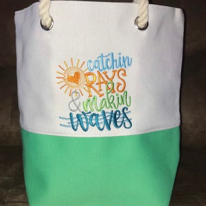 May include: White canvas tote bag with a green bottom. The bag has a rope handle and two metal grommets. The bag has an embroidered design of a sun with a heart, and the text "catchin rays & makin waves".