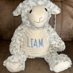 Könnte beinhalten: Ein weißes Plüschlamm mit dem Namen "LIAM" in blauer Stickerei auf der Brust. Das Lamm sitzt auf einem braunen Sofa.