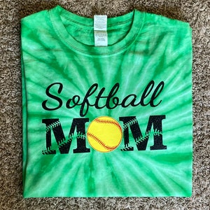 Peut inclure: T-shirt vert tie-dye avec les mots "Softball MOM" en écriture noire et en lettres majuscules. La lettre "M" est ornée de coutures de baseball et d'une balle de softball jaune au centre.