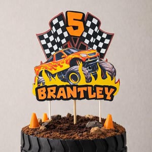 Puede incluir: Una tarta de cumplea&ntilde;os con tem&aacute;tica de monster truck con un dise&ntilde;o de neum&aacute;tico negro. La tarta est&aacute; decorada con monster trucks de juguete, conos de tr&aacute;fico naranjas y un adorno que dice "Brantley" y "5".