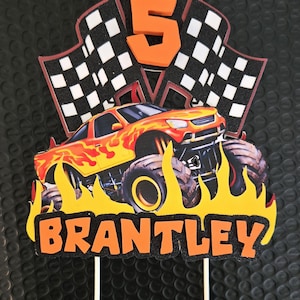 Puede incluir: Adorno de pastel colorido con un cami&oacute;n monstruo con llamas, el n&uacute;mero 5 y banderas a cuadros. El nombre "BRANTLEY" se muestra en letras naranjas. Ideal para una celebraci&oacute;n de cumplea&ntilde;os.